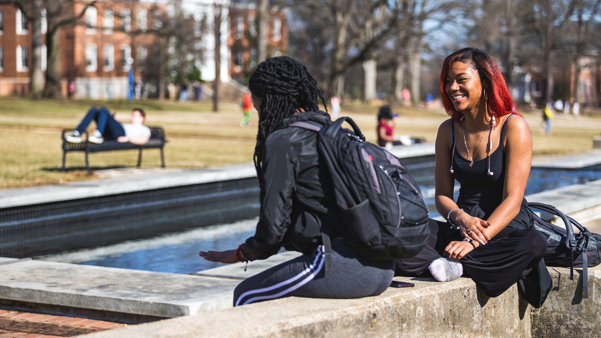 Two students sit on the edge of a water feature outdoors on the Mall on an unseasonably warm February day.