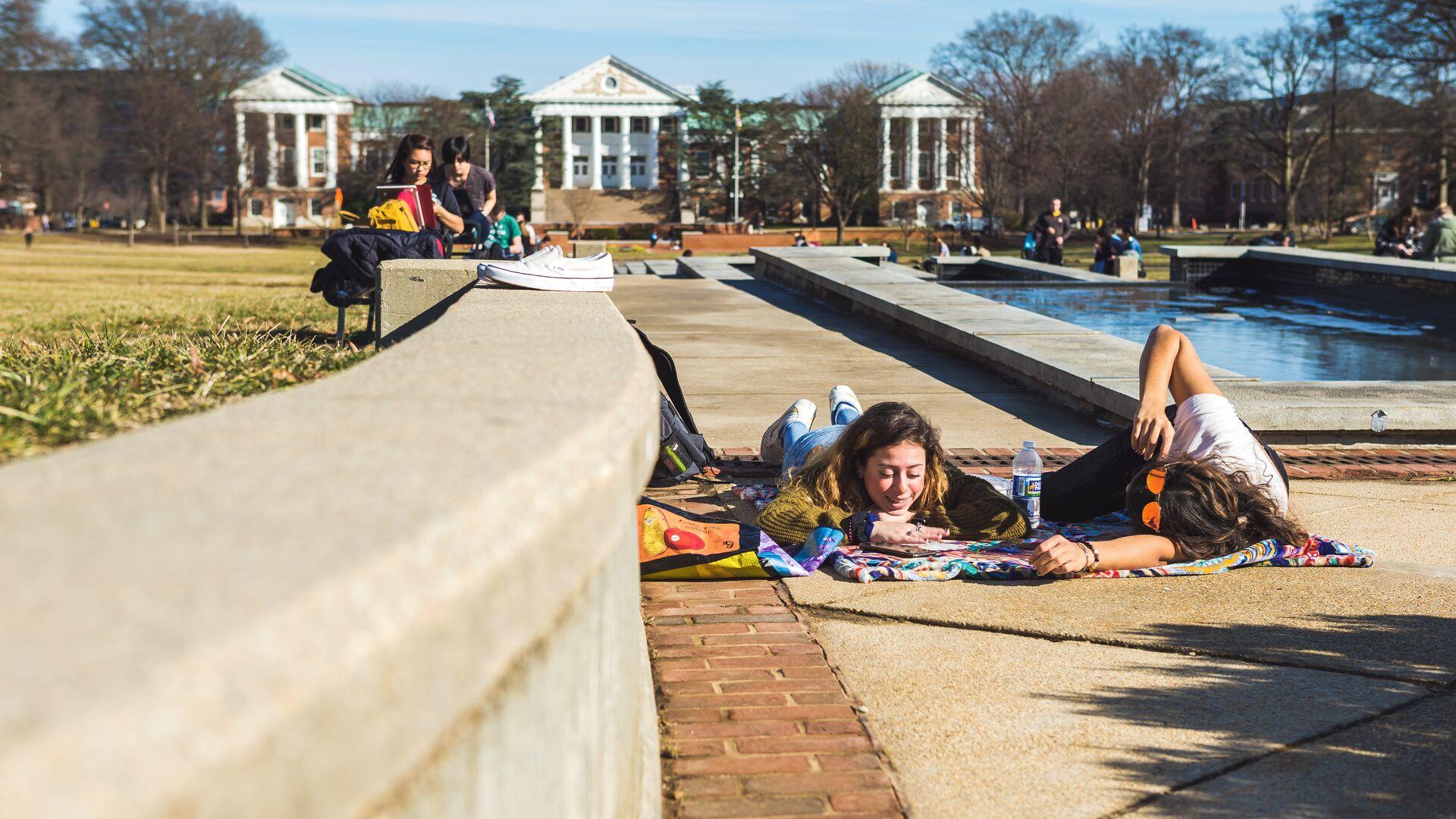 Two female students lie on a blanket outdoors on the Mall on an unseasonably warm February day.
