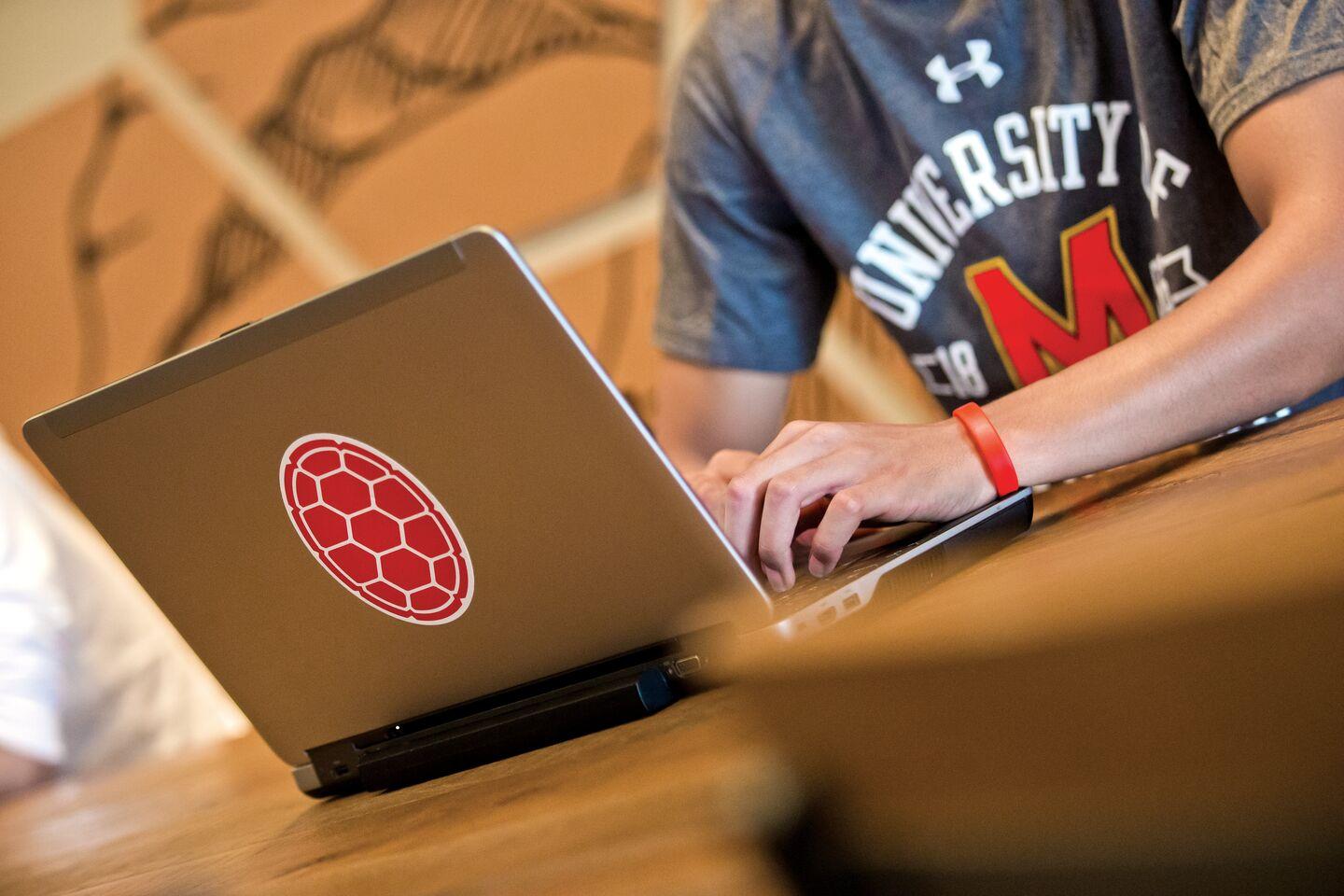 A picture of a male student using his laptop on a table in Starbucks with another student in the background, shell sticker on the laptop.